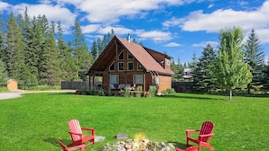 Une cabane en bois avec un toit en pente, une terrasse et un foyer entouré de chaises Adirondack rouges.