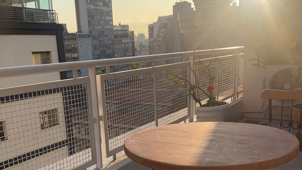 Un balcón con una mesa redonda y dos sillas, barandilla metálica y vistas de edificios de la ciudad al fondo.