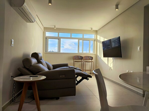 A modern living room with a sofa, a dining table, and a TV mounted on the wall.