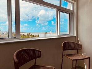 A room with a large window overlooking a beach, two wicker chairs, and a white wall.