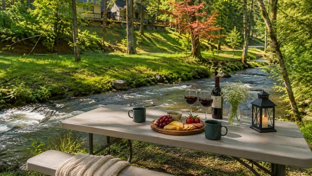 A picnic setup by a river with a table, wine glasses, a bottle, and food.