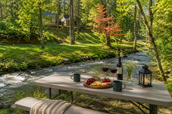 A picnic setup by a river with a table, wine glasses, a bottle, and food.