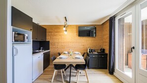 A compact kitchen with a dining area, wooden walls, a flat-screen TV, and a sliding glass door to a balcony.