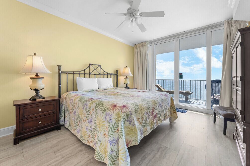 A bedroom with a bed, two bedside lamps, a ceiling fan, a balcony with a view, and a nightstand.