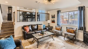 A spacious living room with a dark leather sofa, patterned armchairs, a coffee table, and a large window with curtains. - Spacious Home Near Tennyson & Olde Towne - Sauna + Massage Chair + WiFi (Arvada)