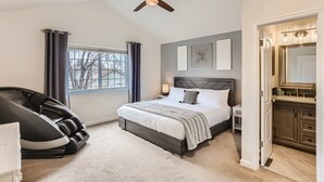 A bedroom with a bed, a massage chair, a ceiling fan, a window with curtains, a nightstand, and a bathroom with a mirror and sink. - Spacious Home Near Tennyson & Olde Towne - Sauna + Massage Chair + WiFi (Arvada)