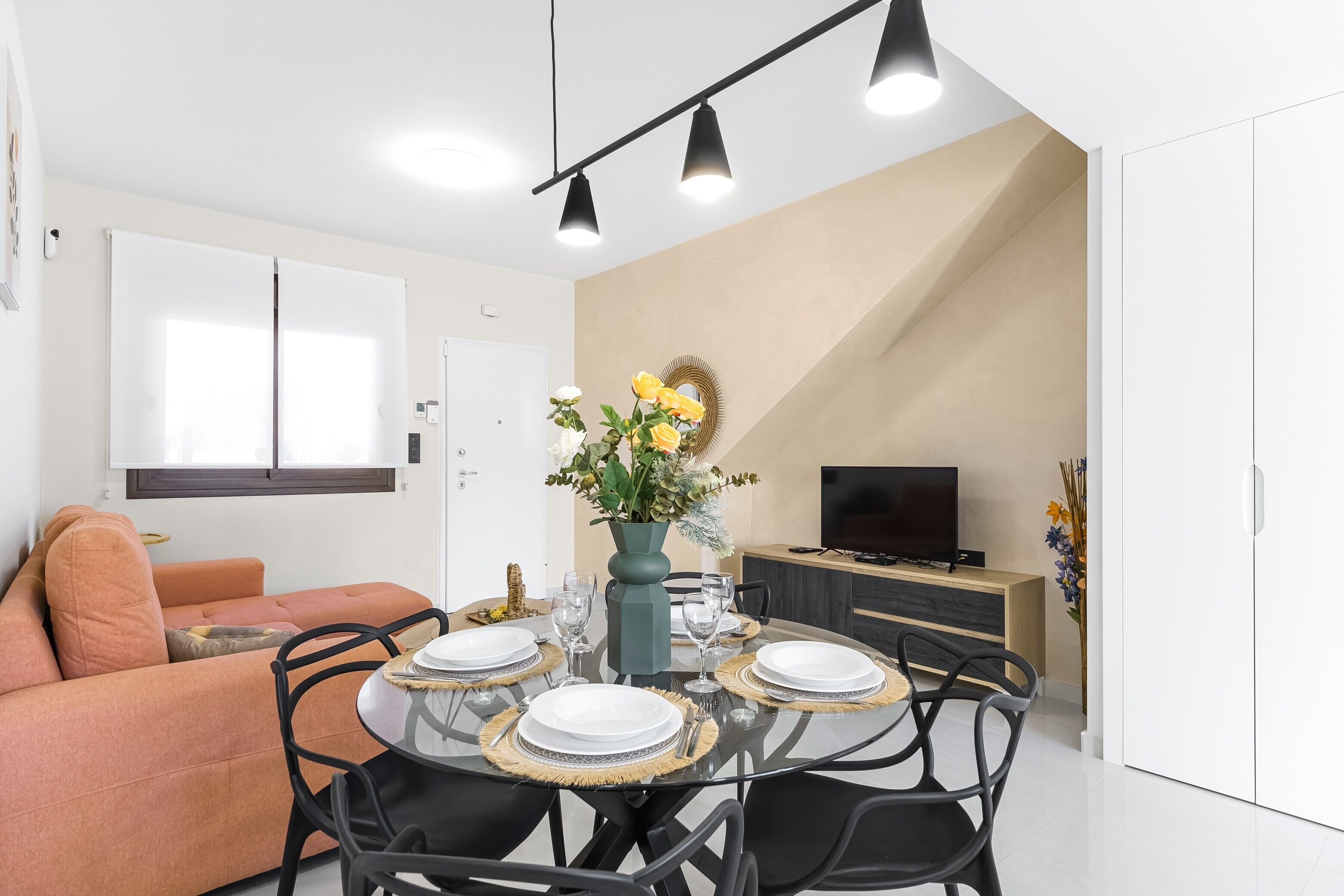 A modern living room with a sofa, dining table set for four, a TV, and a staircase leading to an upper level.
