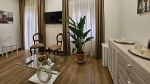 Ein modernes Wohnzimmer mit einem Glastisch, zwei Holzstühlen, einer Topfpflanze, einem Flachbildfernseher und einem Essbereich mit einem weißen Tisch und Tellern.