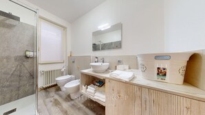 Moderni kylpyhuone, jossa on lasinen suihkuseinä, valkoinen wc-istuin, peilillä varustettu pesuallas ja puinen allaskomero, jonka päällä on pyyhkeitä.