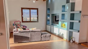 Una sala de estar moderna con un sofá seccional gris, un televisor montado en la pared y una ventana con vista a la ciudad.