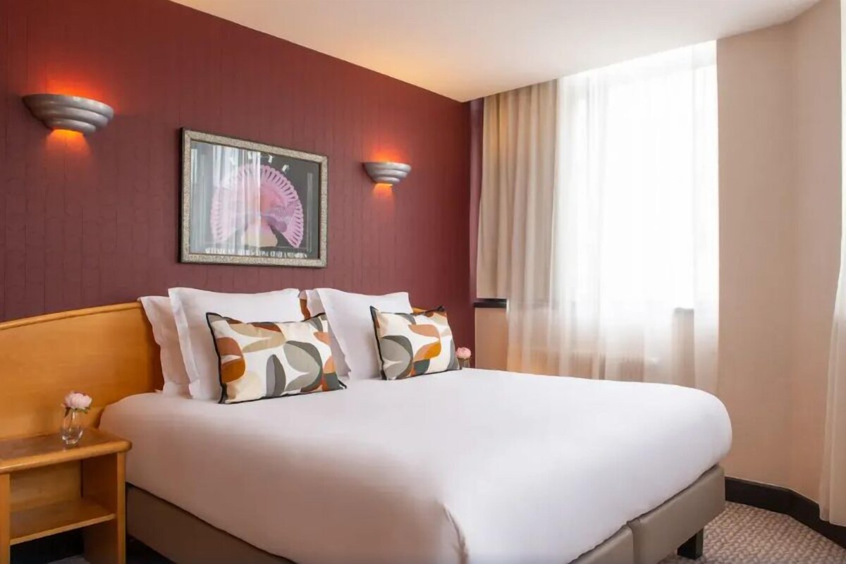 Une chambre d’hôtel avec un grand lit, deux appliques murales, un tableau encadré et une fenêtre avec des rideaux.