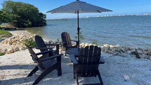 A beachside seating area with Adirondack chairs and a large umbrella, overlooking a calm sea and distant cityscape.