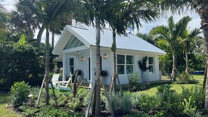A small, single-story house with a white exterior and a metal roof, surrounded by lush greenery and palm trees.