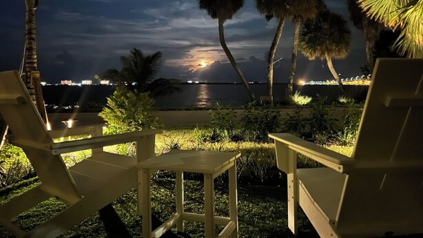 A nighttime view of a beach with lounge chairs and palm trees, overlooking a body of water with city lights in the distance.