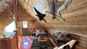 L’intérieur d’une cabane en rondins présente un plafond haut, des murs en bois et un espace de vie chaleureux avec un canapé, des fauteuils et une table basse.