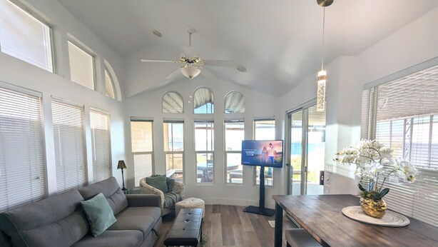Un salón amplio con un sofá gris, una mesita de centro y un televisor. Hay ventanas grandes y un ventilador de techo.