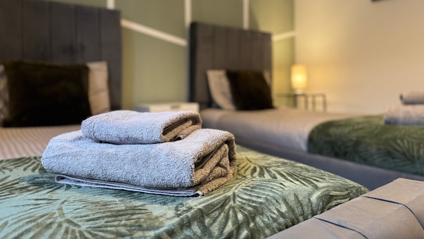 A hotel room with two beds, a green patterned bedspread, and a folded towel on the left bed.
