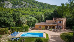 Une maison à deux étages avec une piscine, entourée d’une végétation luxuriante et de collines.