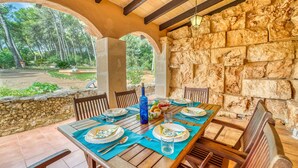 Un espace de restauration extérieur abrité, doté de murs en pierre et de poutres en bois, avec une table dressée pour deux personnes, comprenant des assiettes, des couverts et une bouteille d’eau.