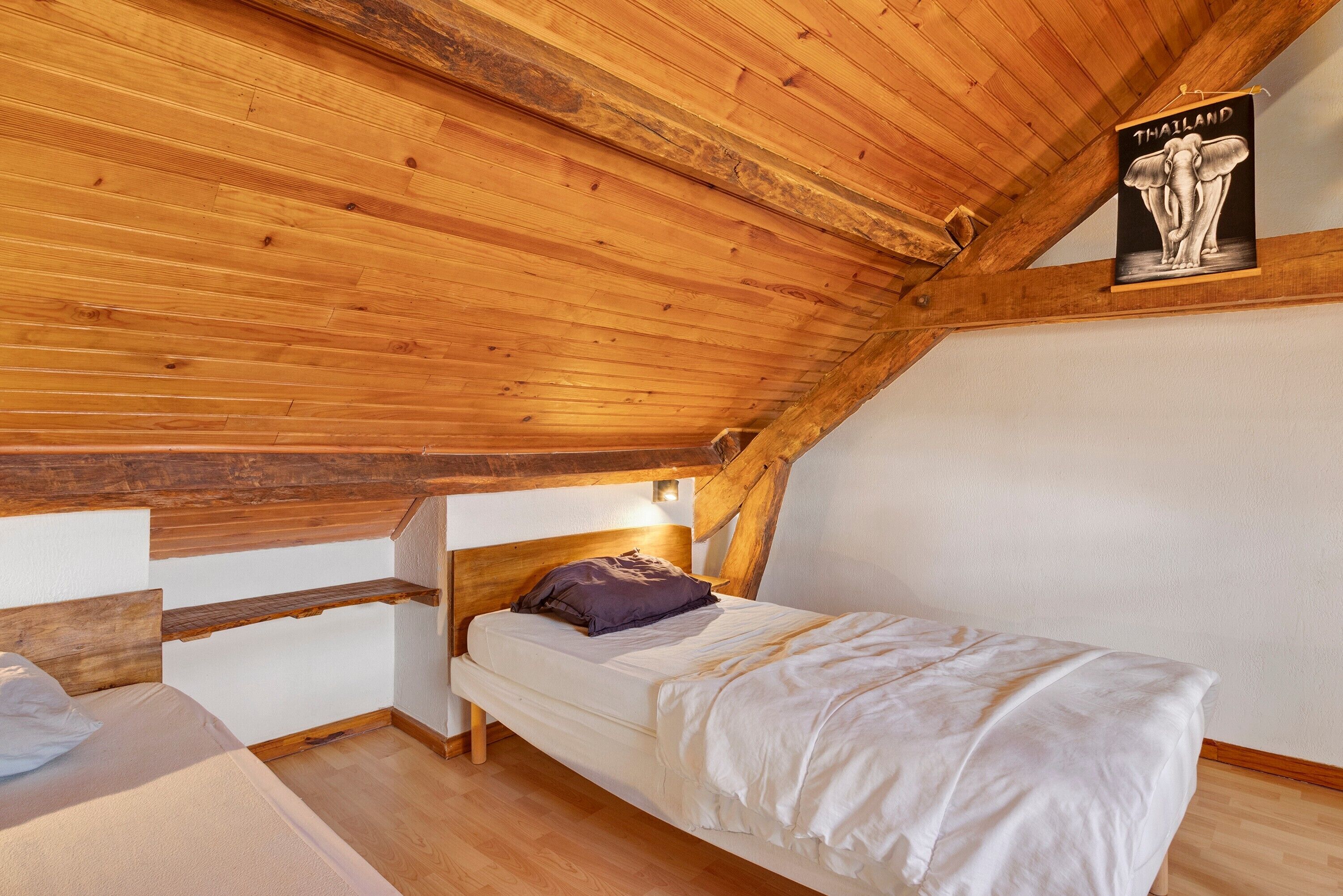 Une chambre avec un plafond en pente, des poutres en bois, un lit simple avec un oreiller et une affiche d’un éléphant accrochée au mur.
