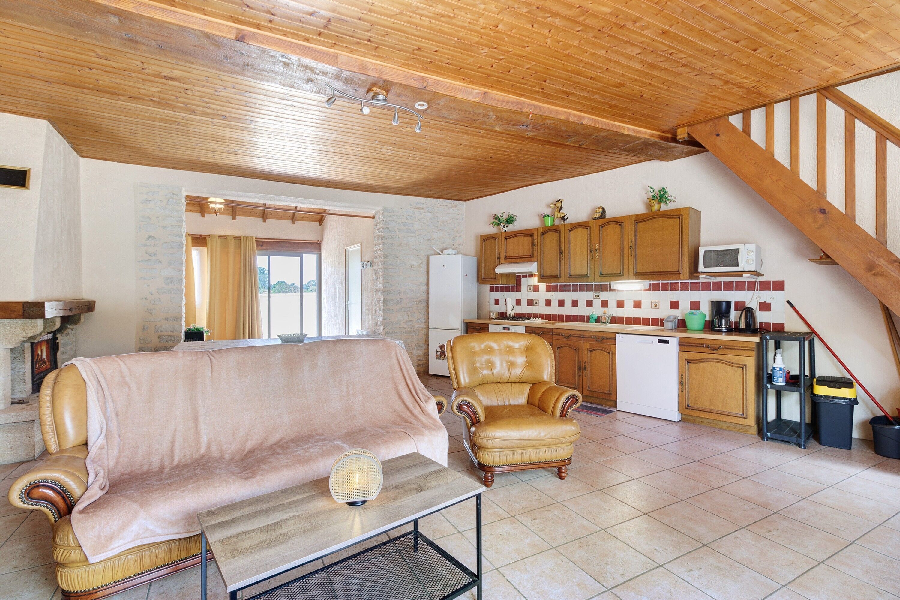 Ein gemütliches Wohnzimmer mit einer Couch, einem Sessel und einem kleinen Tisch. Es gibt eine Küchenzeile mit Holzschränken und einer Mikrowelle.