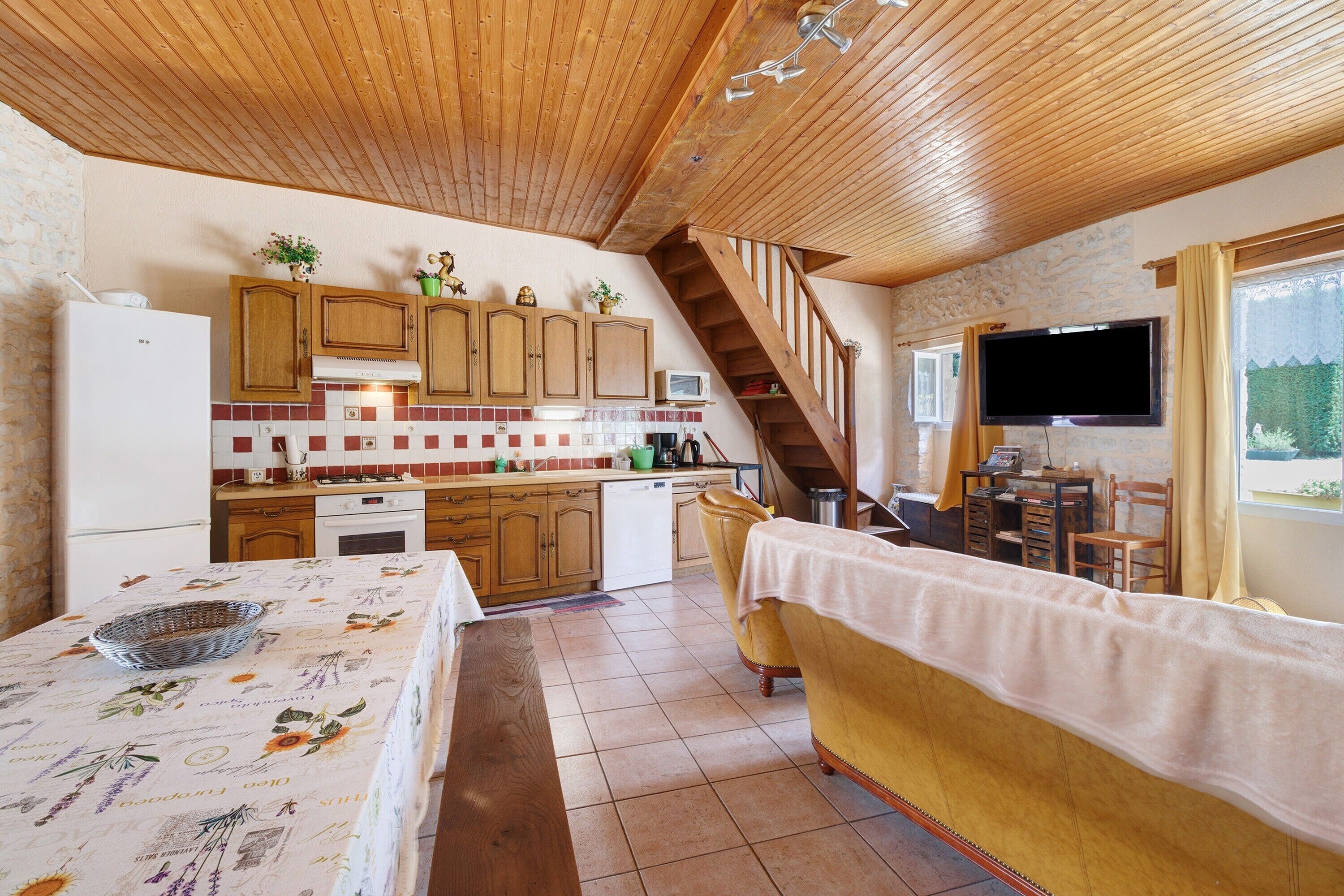 A kitchen with wooden cabinets, a dining area with a table and chairs, a television, and a staircase.