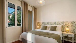 Een slaapkamer met een bed, gordijnen, een nachtkastje met een lamp, een raam en een wand met bloemenprint.