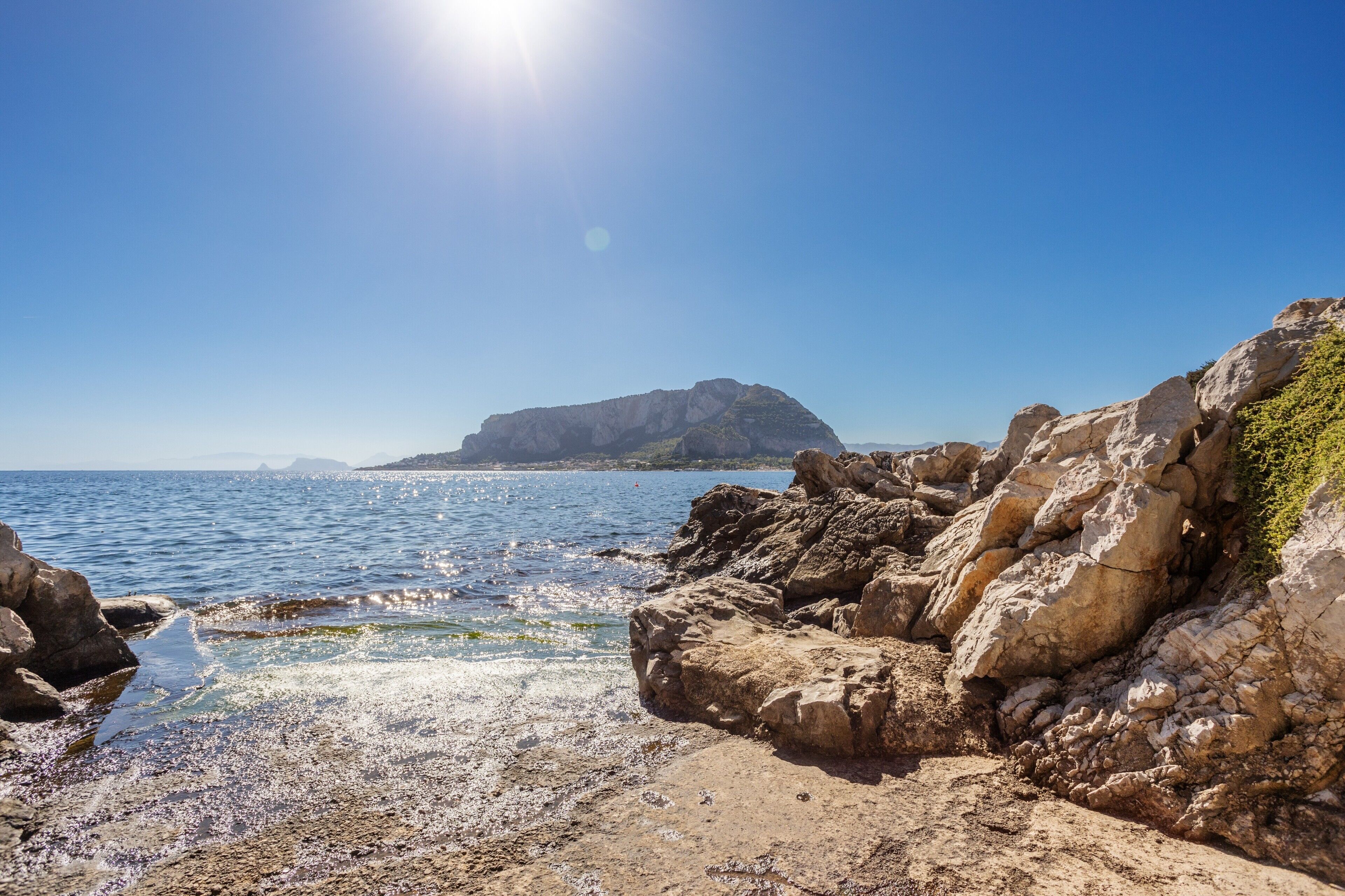 Una costa rocciosa con acque limpide, una montagna in lontananza e un sole splendente.