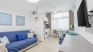 A modern living room with a blue sofa, a dining area, a kitchen with a washing machine, and a flat-screen TV mounted on the wall. - Studio 'Flat Blu Palermo Mondello' with Shared Pool, Shared Terrace & Air Conditioning (Palermo)