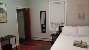 A bedroom with a bed, a bedside table, a mirror, a window with blinds, a door, and a small desk with a chair. - The Town-Suite at Wanskuck NEW 2-BR APT  Parking (Povidence)