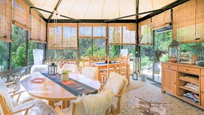 Une pièce avec une table ronde en bois, des chaises recouvertes de coussins en fausse fourrure blanche, une grande fenêtre avec des stores en bambou et une vue sur la verdure à l’extérieur.