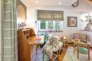 A cozy living room with a wooden desk, a comfortable chair, a small table with a lamp, and a window with a view of greenery.