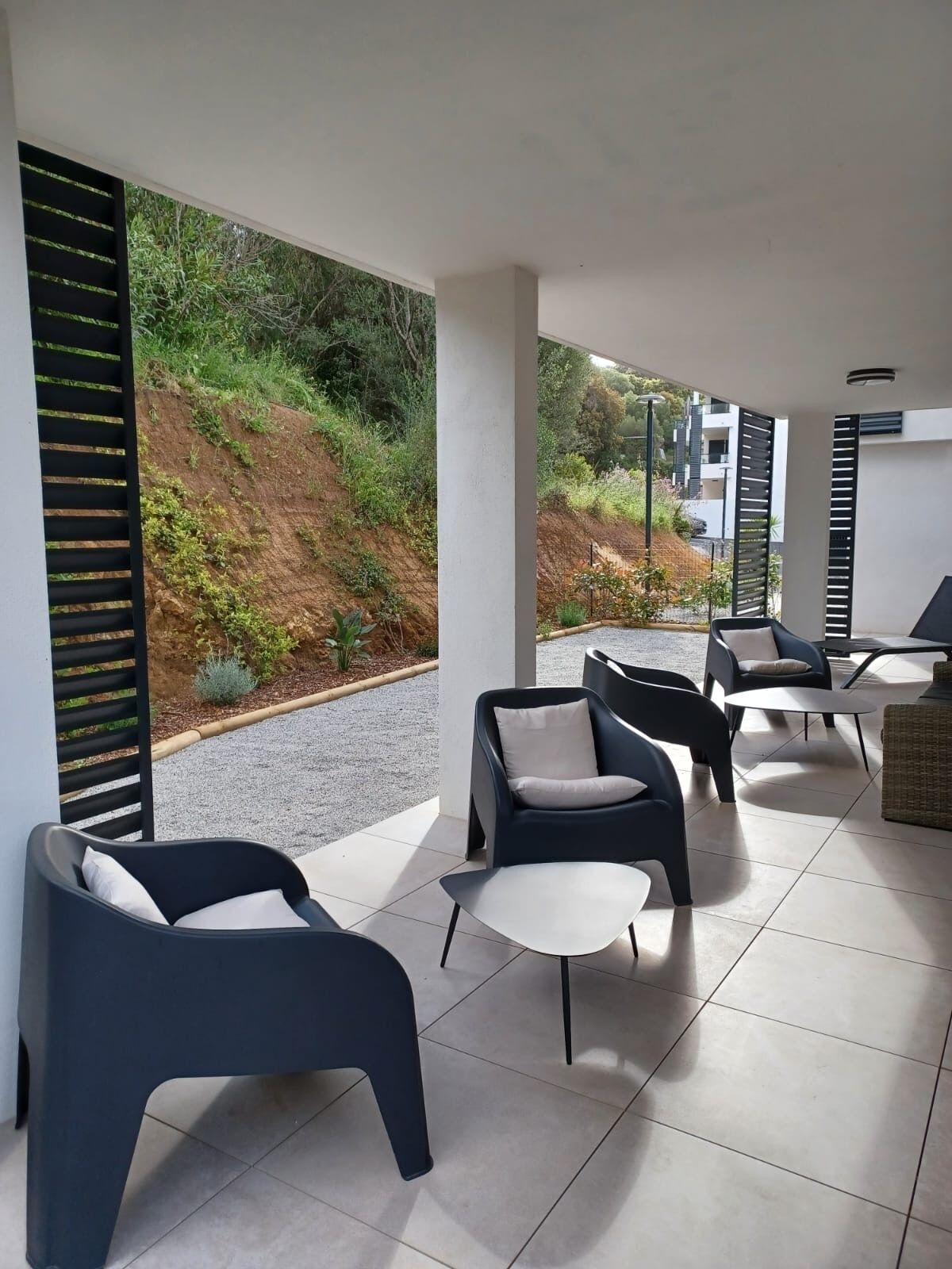 Uma área de estar moderna ao ar livre, com cadeiras contemporâneas e uma pequena mesa, com vista para um jardim paisagístico e um edifício ao fundo.
