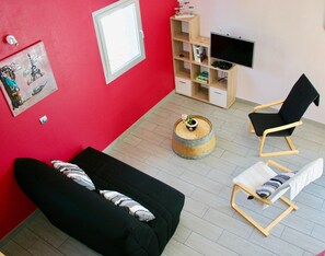 A room with a black sofa, a wooden chair, a small round table with a flower, a TV on a wooden stand, and a bookshelf.