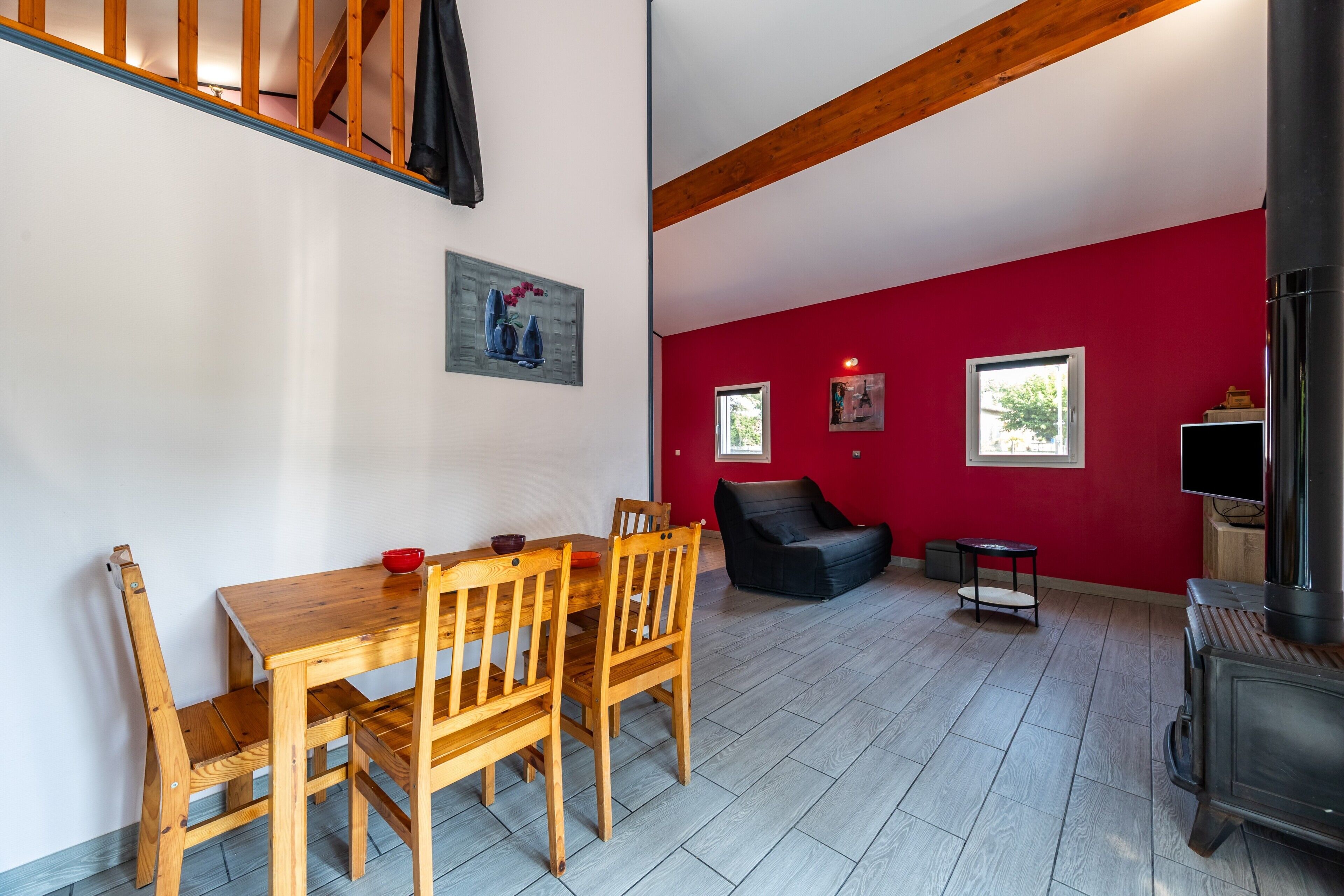 Un comedor con mesa y sillas de madera, pared roja con dos ventanas y un sofá negro.