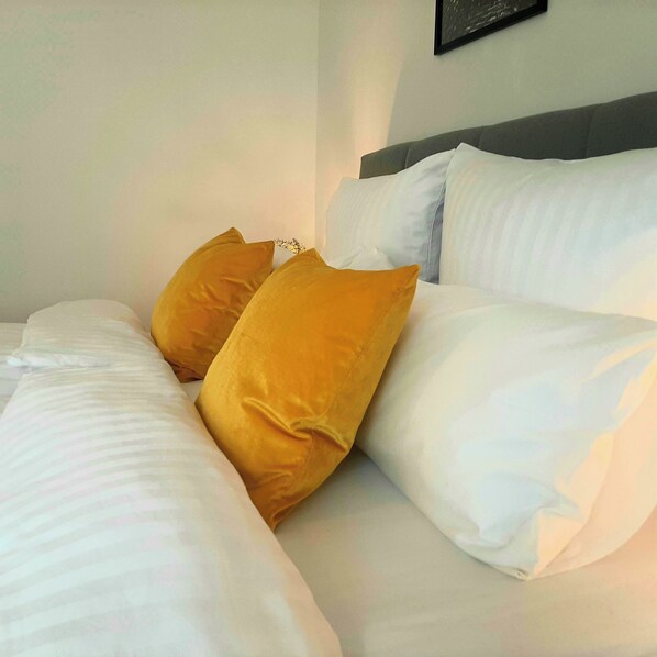 A neatly made bed with white and mustard yellow pillows, a grey headboard, and a framed picture on the wall.