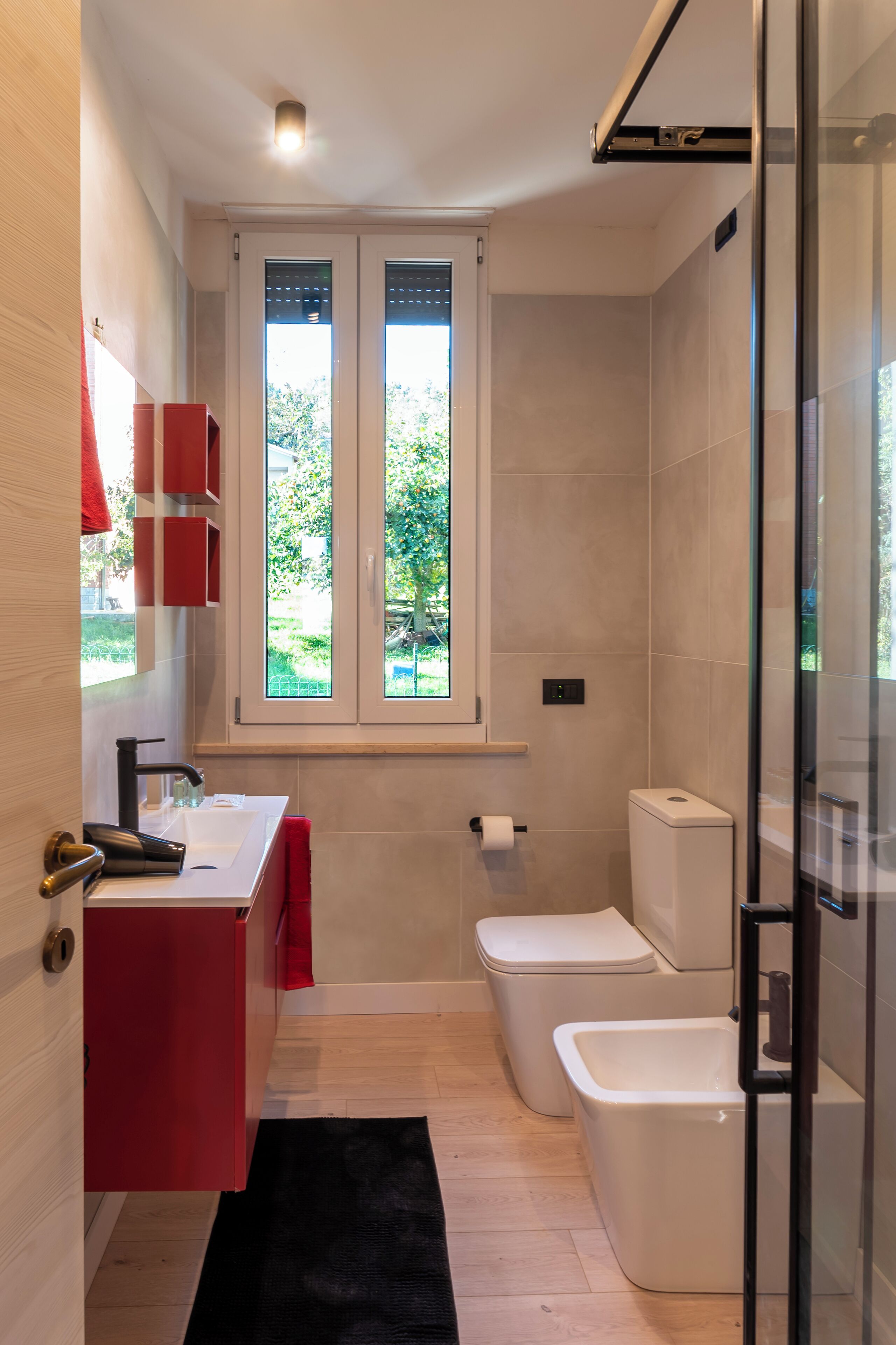 Une petite salle de bain avec un lavabo blanc, des meubles rouges, une cabine de douche en verre et une fenêtre donnant sur un espace verdoyant.
