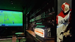A bar area with a television displaying a sports game, shelves with bottles, and a mannequin dressed in a FAEM jacket. - IL Corridore - Locanda Bistrò (Medesano)