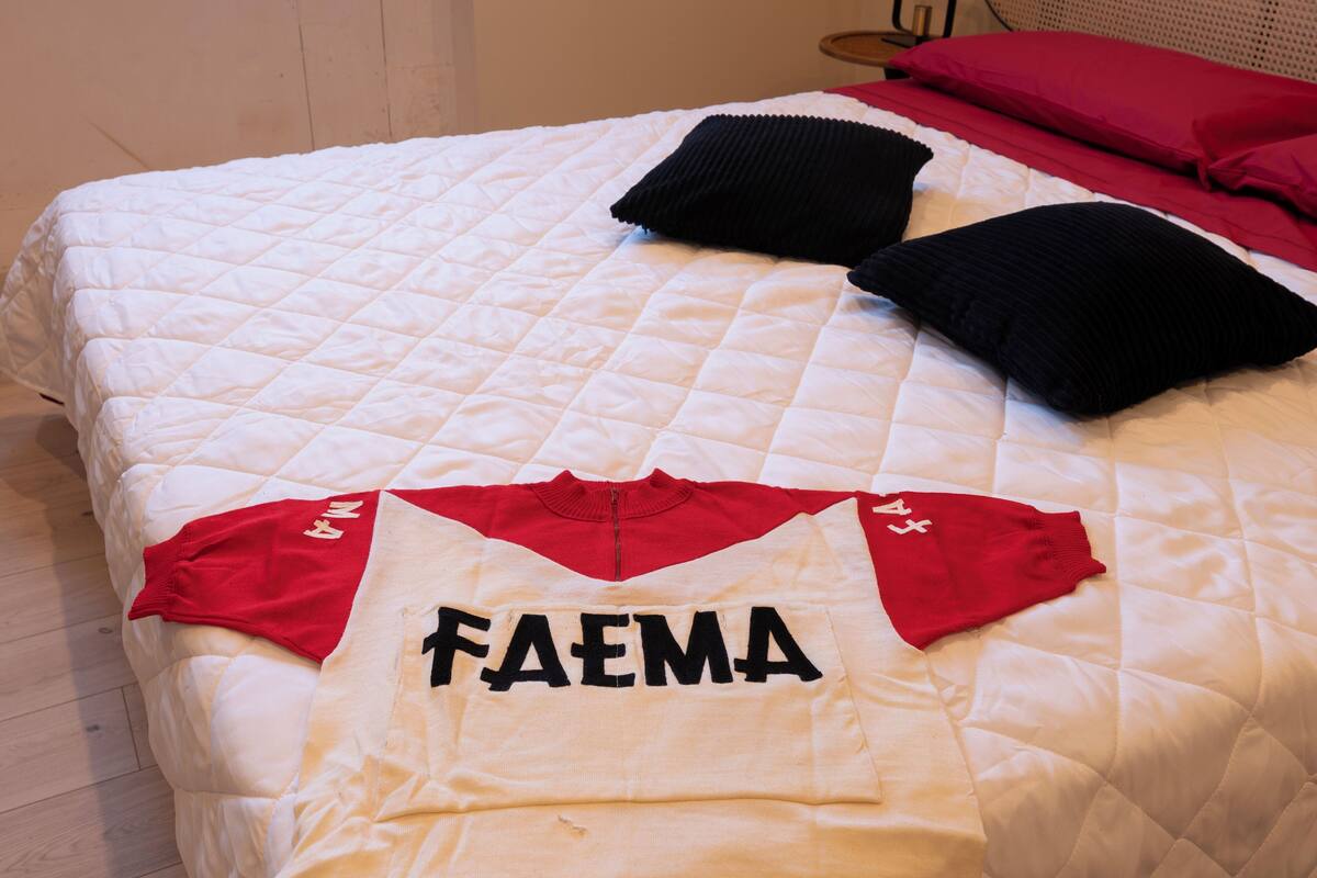 A bed with a quilted white duvet, a red and white FAEMA sports jersey, and two black pillows.