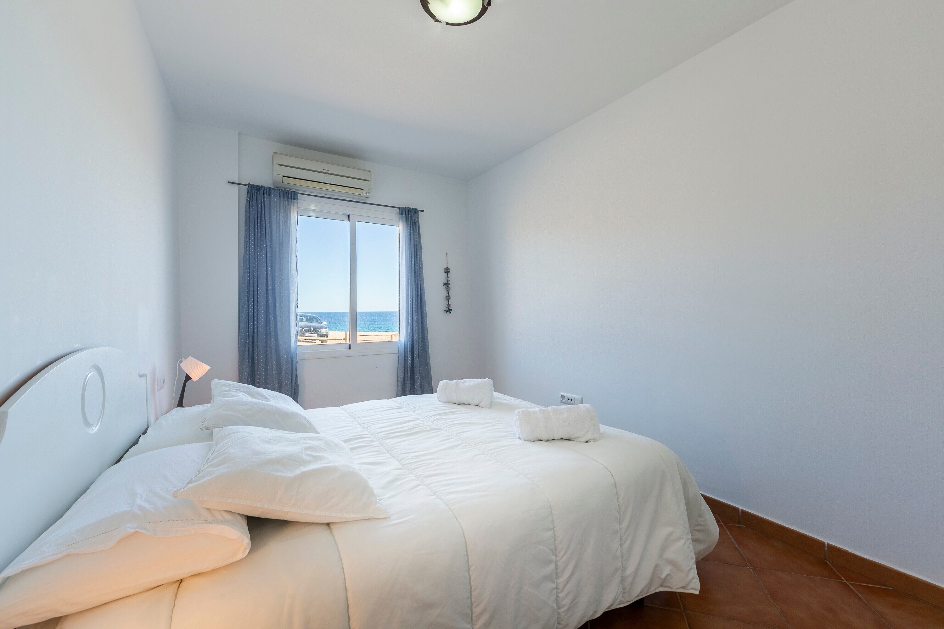 A bedroom with a bed, a window with curtains, an air conditioning unit, and a view of the sea.