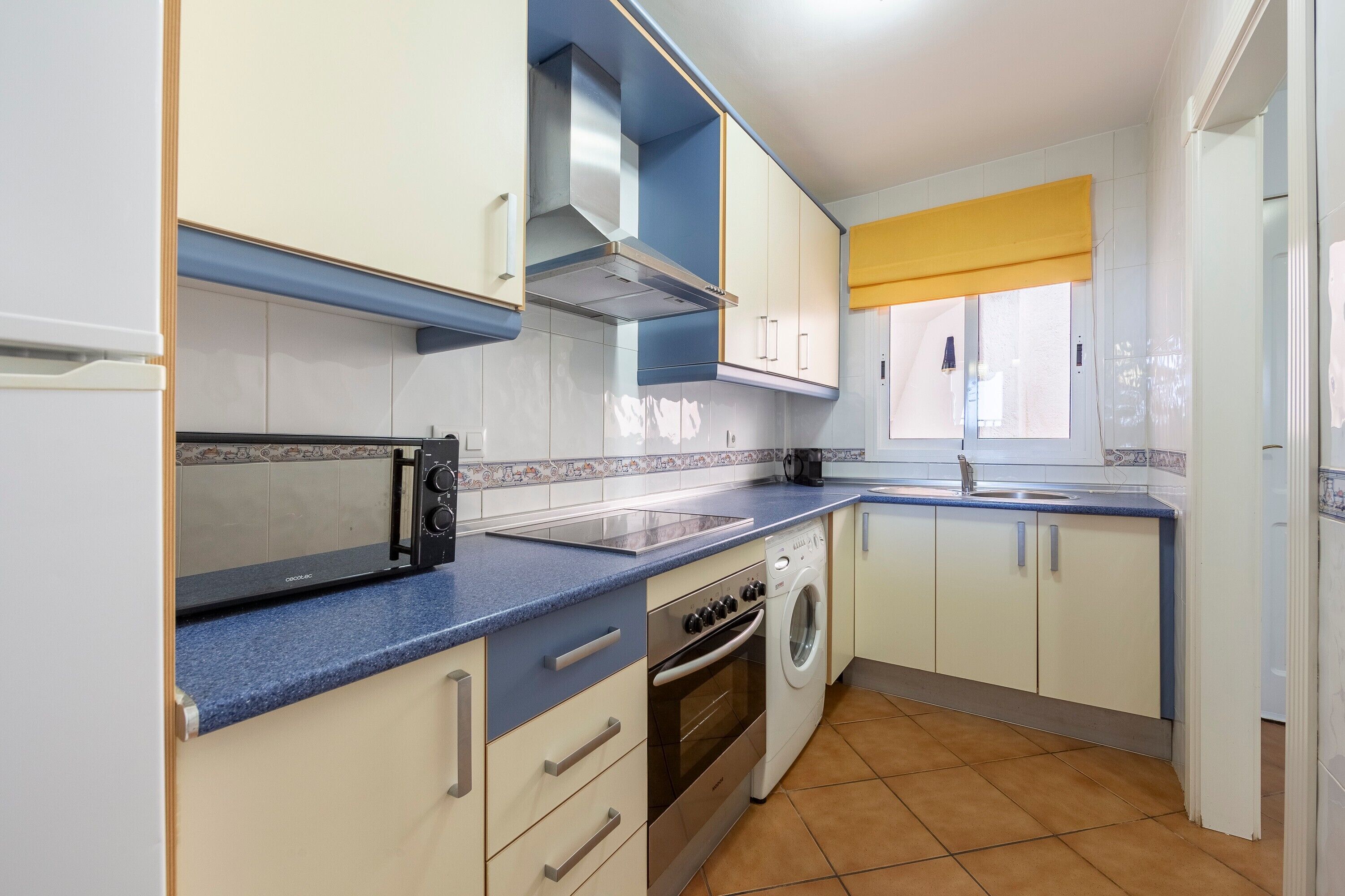 Cocina compacta con microondas, horno y lavadora, con armarios de colores claros y encimera azul.