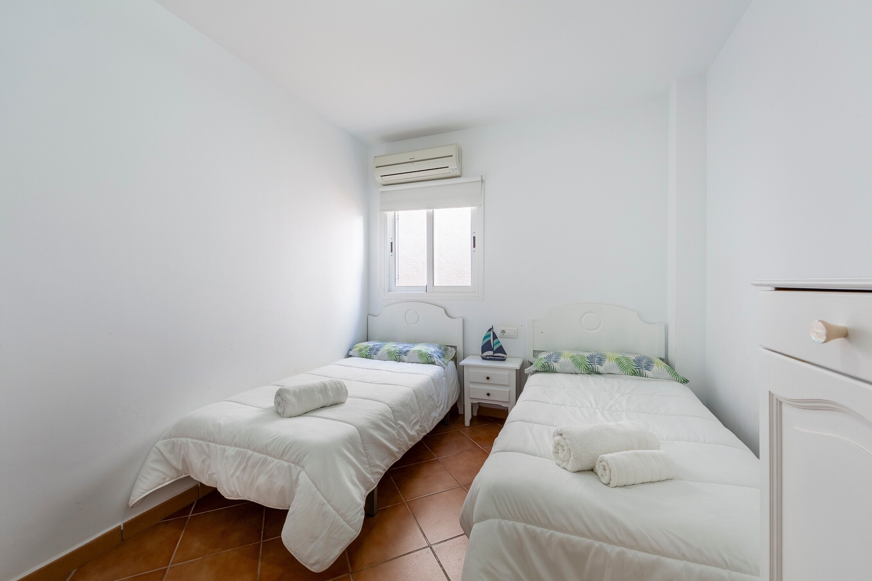 Un dormitorio con dos camas, una ventana, un equipo de aire acondicionado y una mesita de noche con una lámpara.