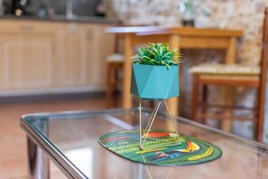 Una mesa de vidrio con un posavasos decorativo, una maceta color turquesa con una planta, y una silla de madera en una habitación con pared de piedra y muebles de madera.