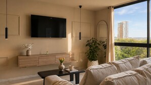 A modern living room with a large TV, a wooden console, a coffee table, a sofa, and a large window with a view of buildings and trees. - CityView 2 BedRoom / Daily cleaning (Guadalajara)
