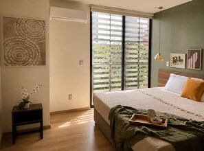 Une chambre à coucher avec un lit, une table de chevet ornée d’un vase, une fenêtre avec des stores et des œuvres d’art accrochées au mur.
