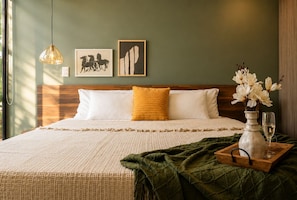 A bed with a wooden headboard, a yellow pillow, and a white pillow. There are two framed pictures on the wall, a hanging light, and a vase with flowers on a tray.