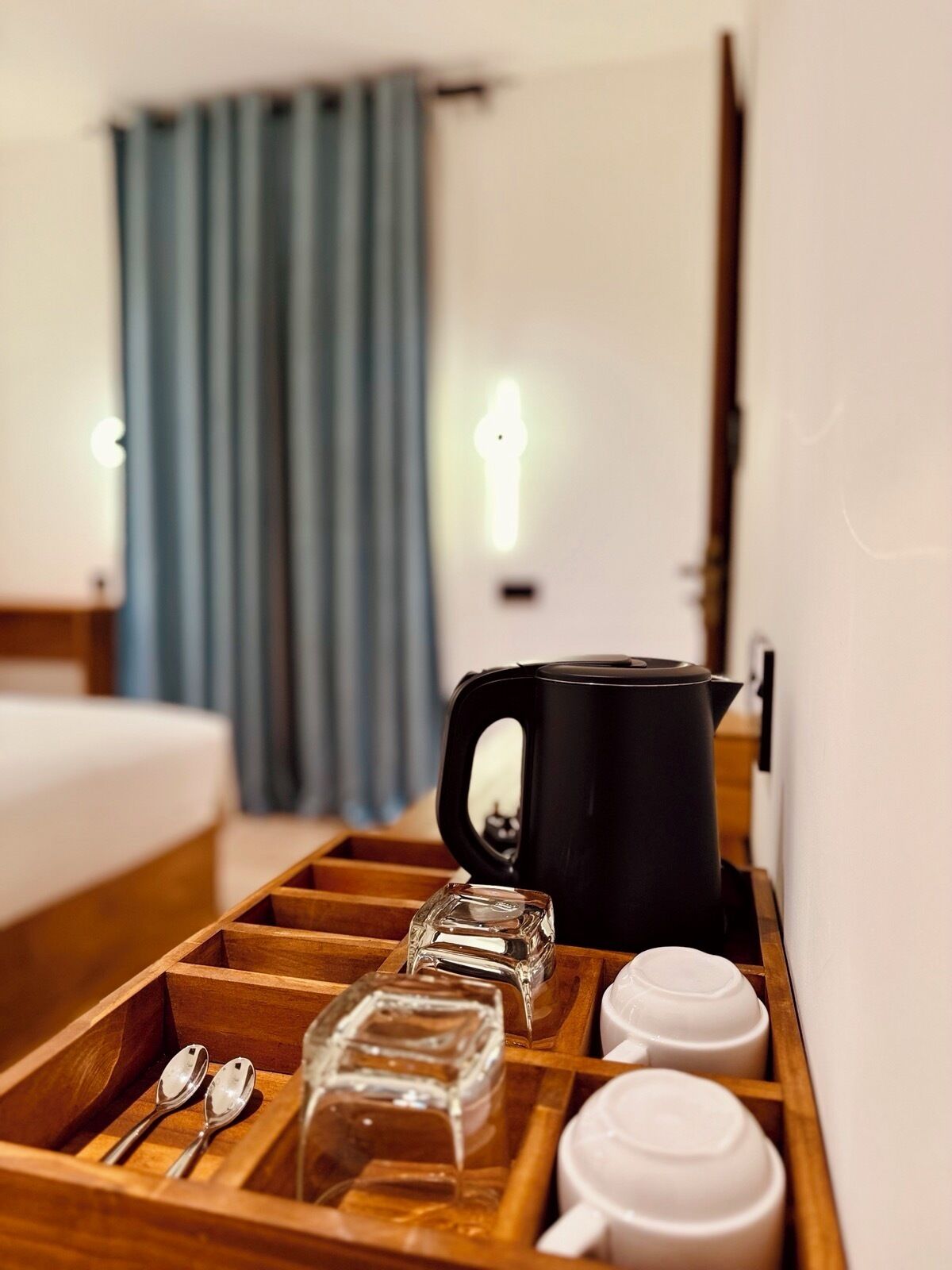 Una camera d'albergo con un vassoio di legno che contiene un decanter di vetro, un bollitore nero, tazze bianche e cucchiaini.