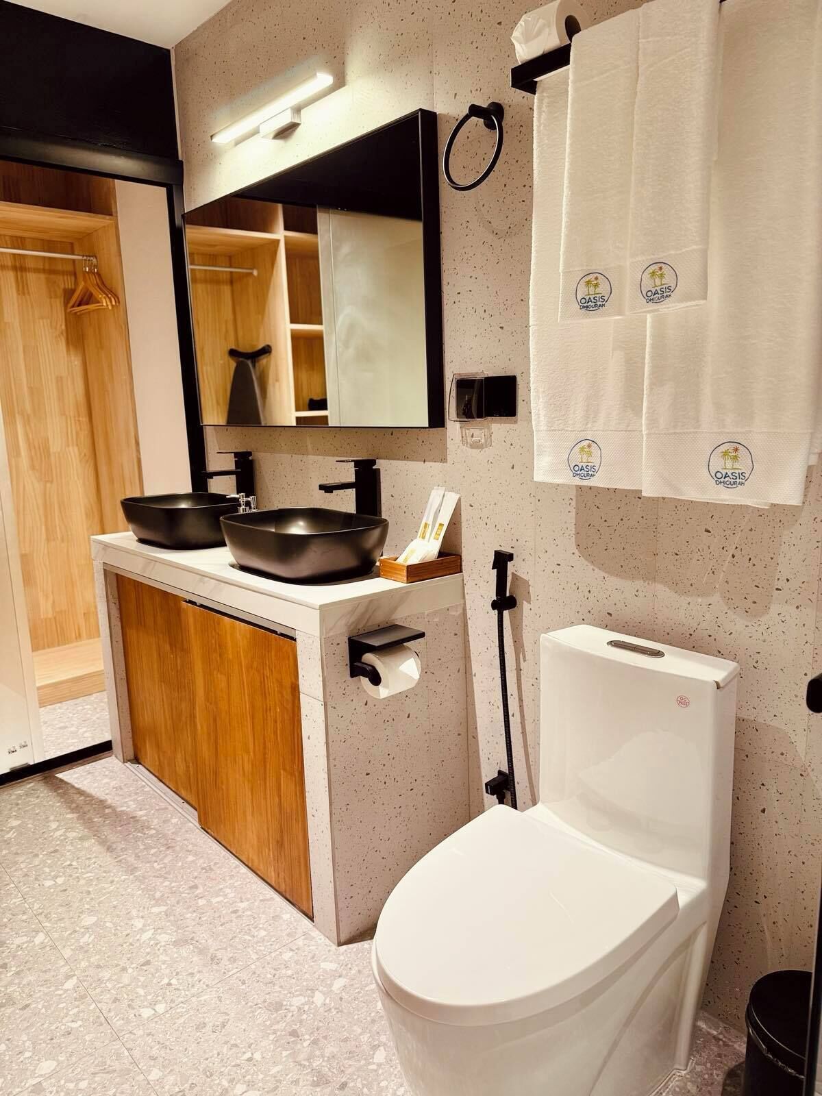 Un baño moderno con un inodoro blanco, un lavamanos negro con forma de cuenco, un espejo sobre el lavamanos y un perchero con toallas.