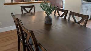 A dining area with a wooden table, chairs, and a vase with plants. There is a ceiling fan above and a kitchen in the background.