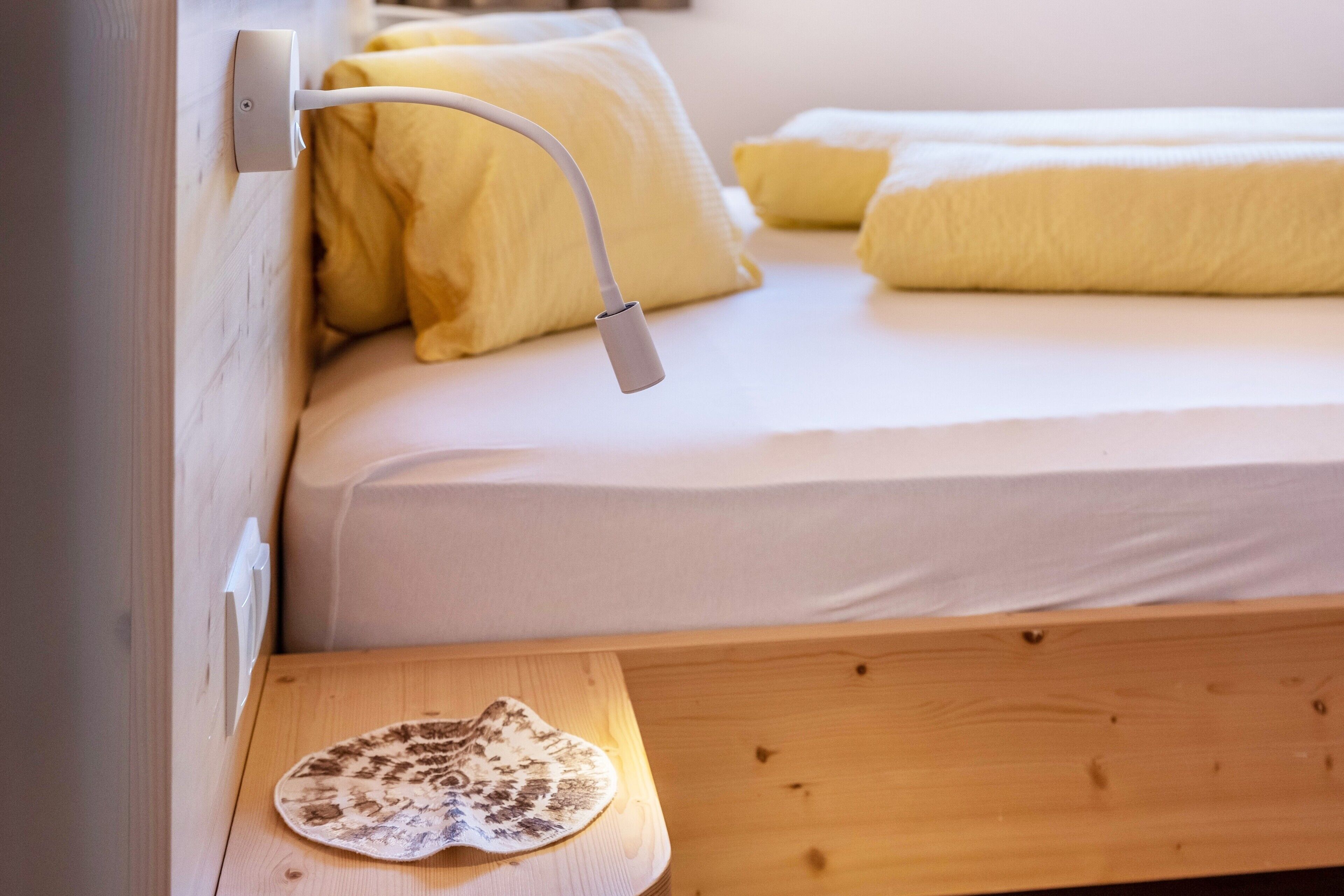 Un lit avec une tête de lit blanche, une table de chevet sur laquelle est posée une nappe décorative, et une lampe fixée au mur.